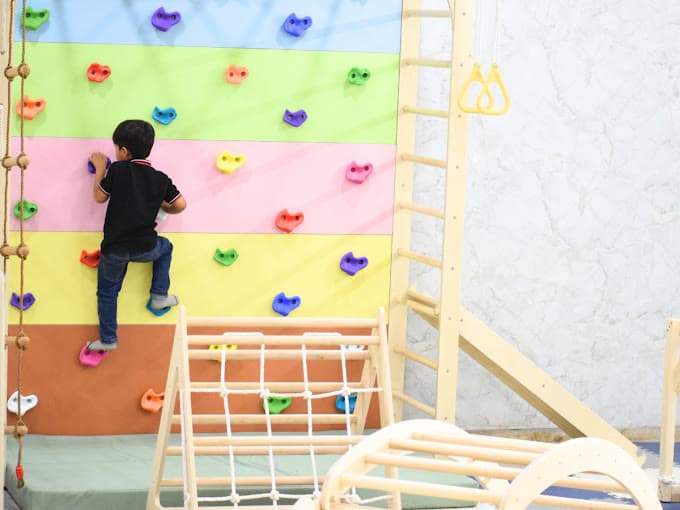 A child climbing a rainbow-coloured indoor climbing wall beside a wooden frame, rings and a rope