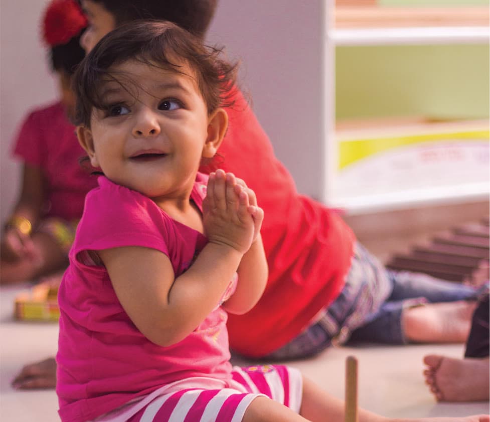 A toddler sitting on a classroom floor, hands together, looking up and away from the camera