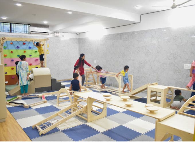 The indoor gym — a teacher guiding children across a wooden balance course, the climbing wall behind