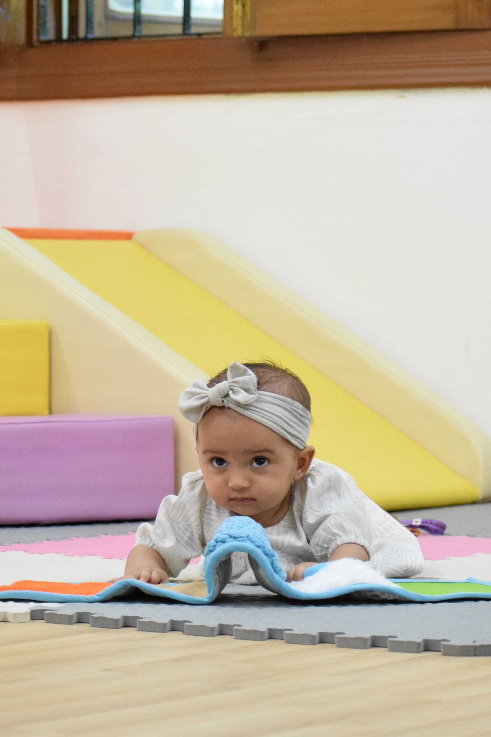 An infant on a play mat in the infant room, a soft-play ramp and wooden pull-along behind her
