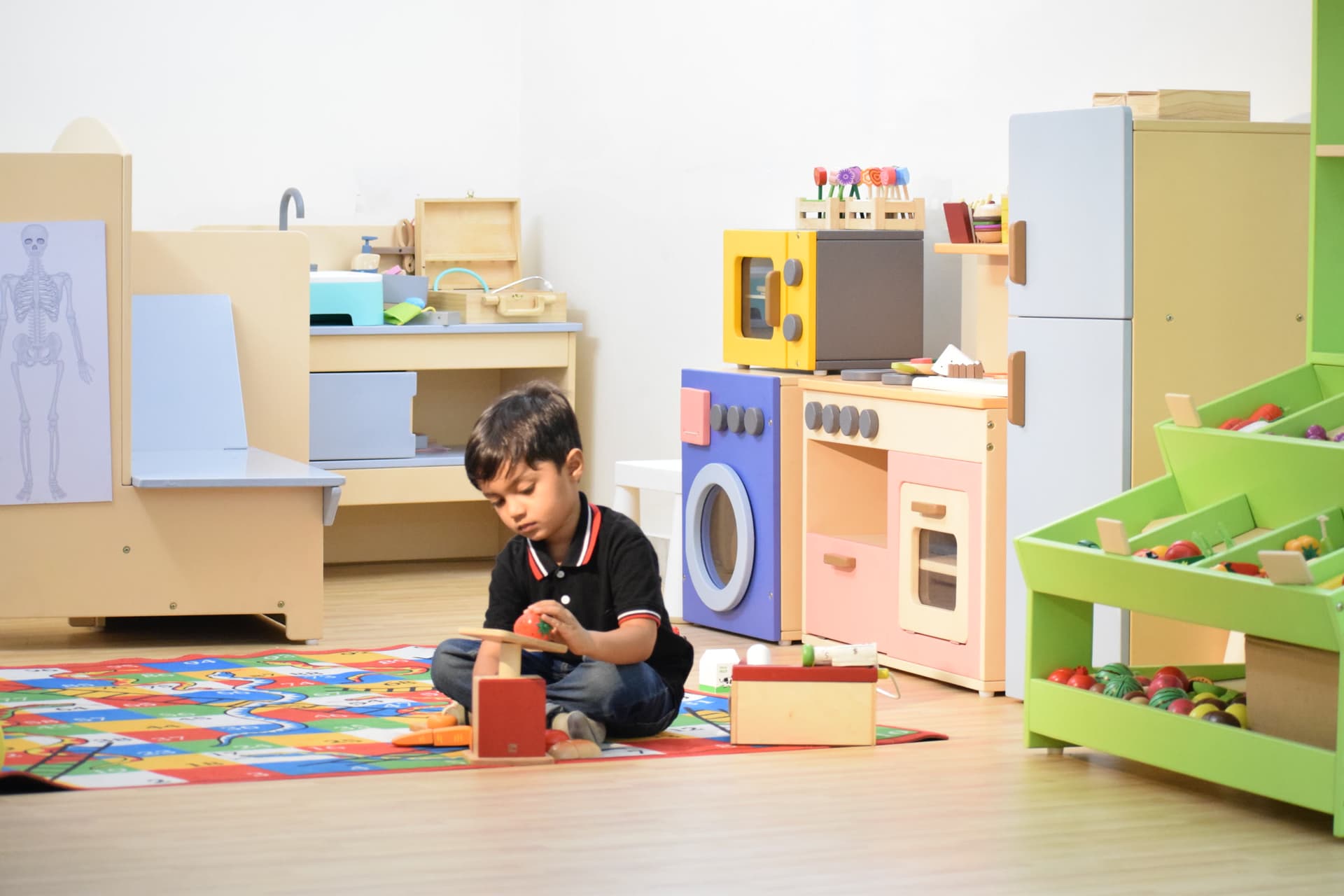 A boy sitting alone on a play mat, absorbed in a wooden toy, in front of a play kitchen and shelves of materials