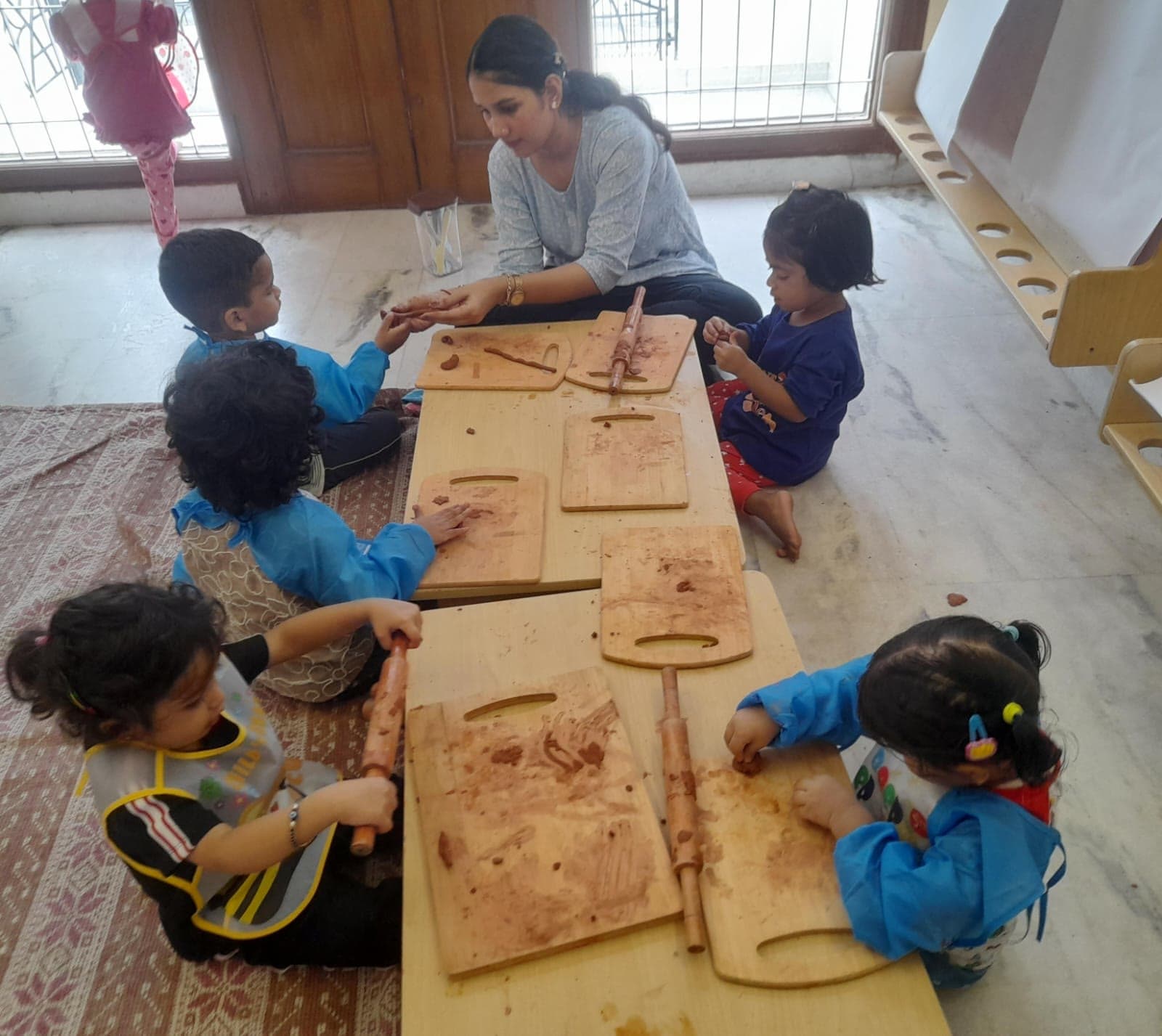 Five children rolling and pressing clay on wooden boards at a low table, a teacher working alongside them