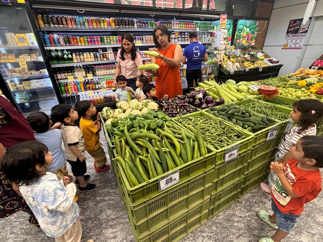 Children at a supermarket produce counter, two teachers holding up a vegetable, crates of greens in front of them