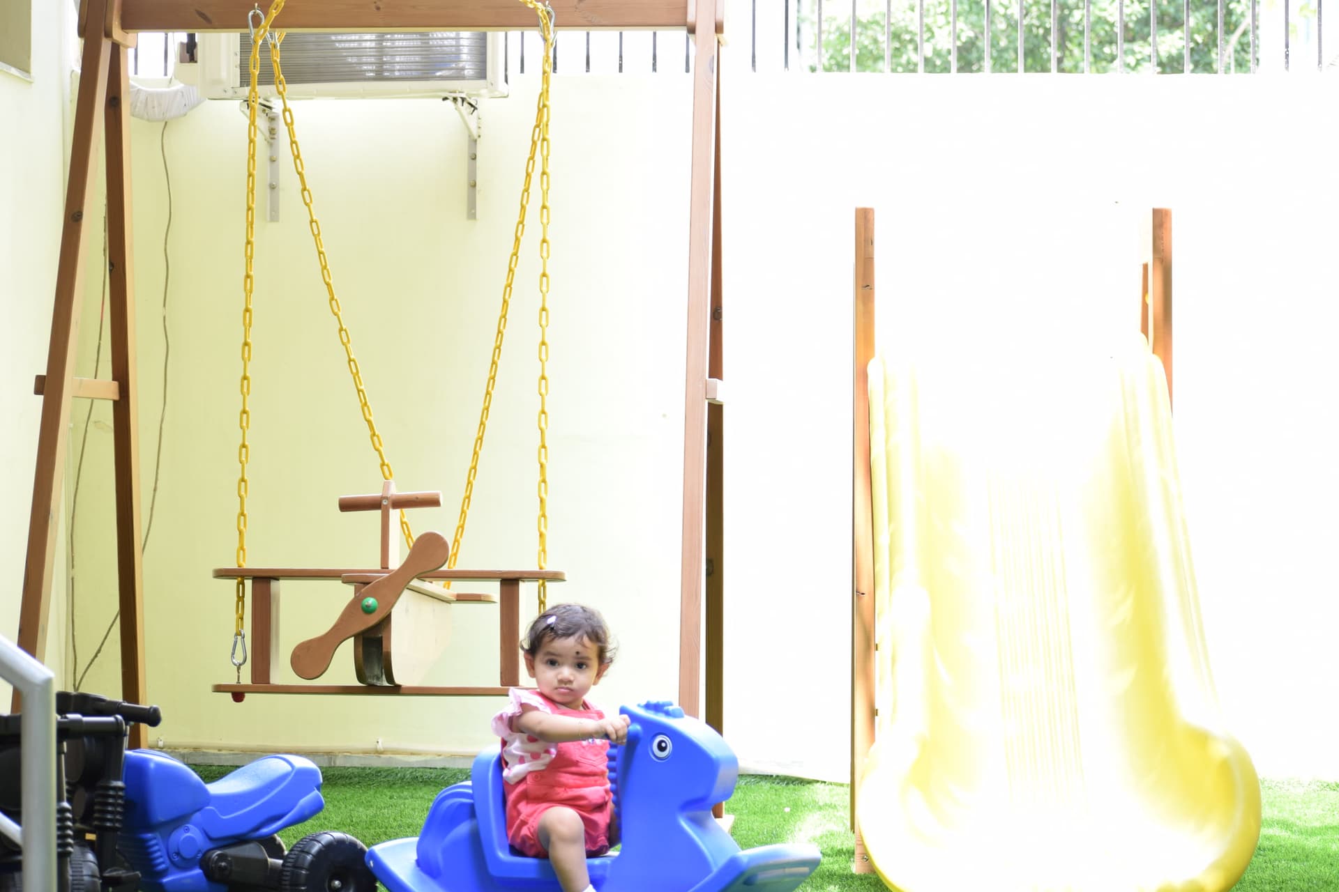 A toddler on a rocking horse on the outdoor play terrace, beside a swing and a slide