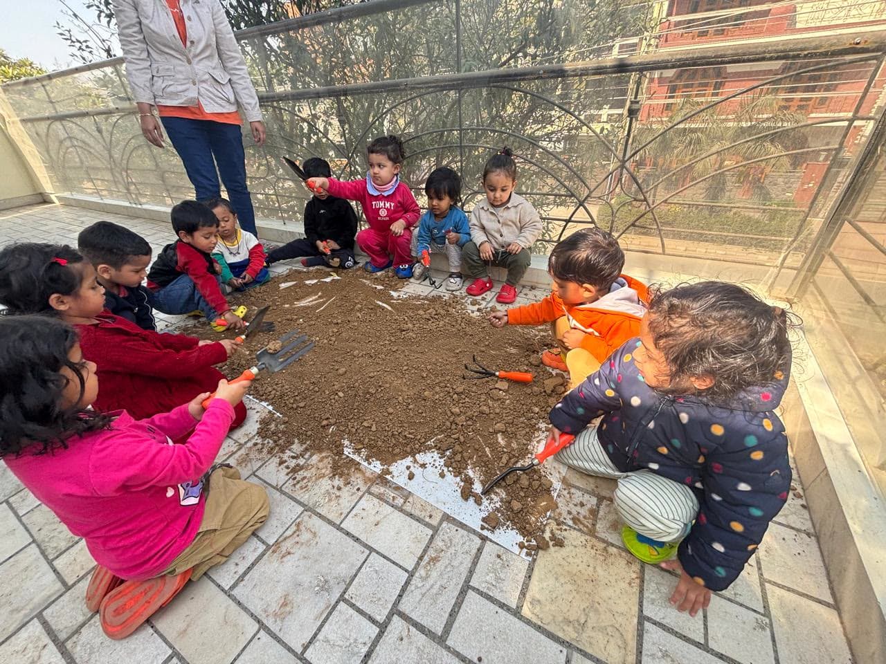 A ring of children turning soil with hand trowels on the terrace, a teacher standing over them