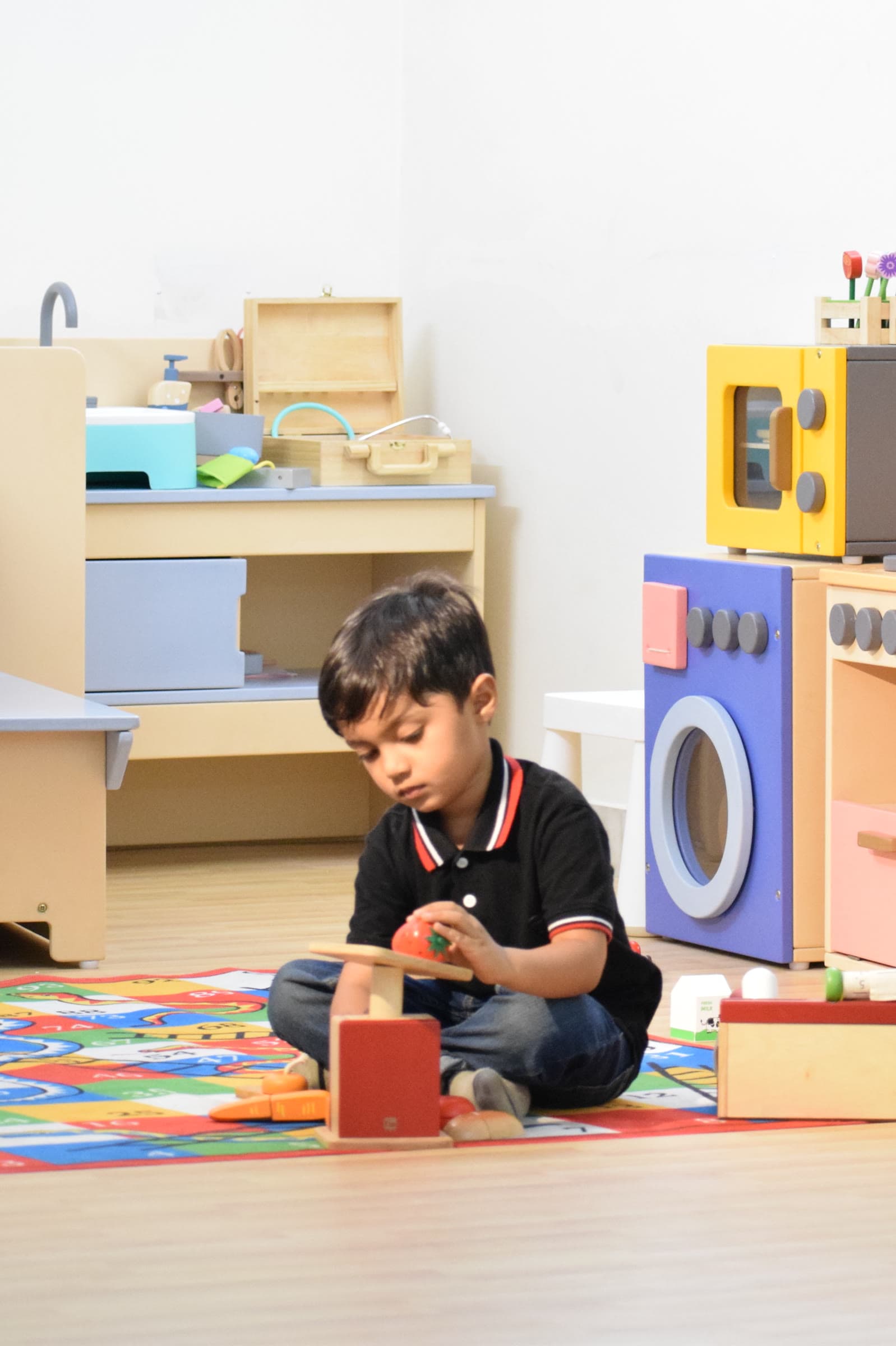A boy sitting on the floor of the preschool room with the play kitchen and shelves of materials behind him