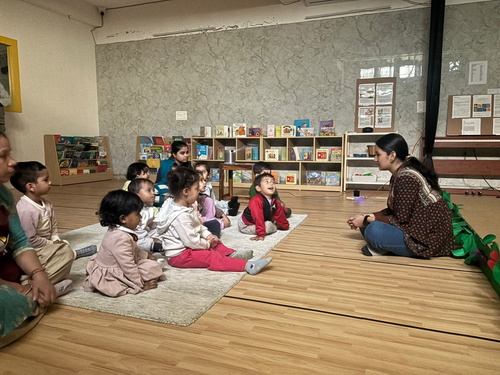 Children sitting on a rug listening to a teacher, shelves of picture books along the wall behind them