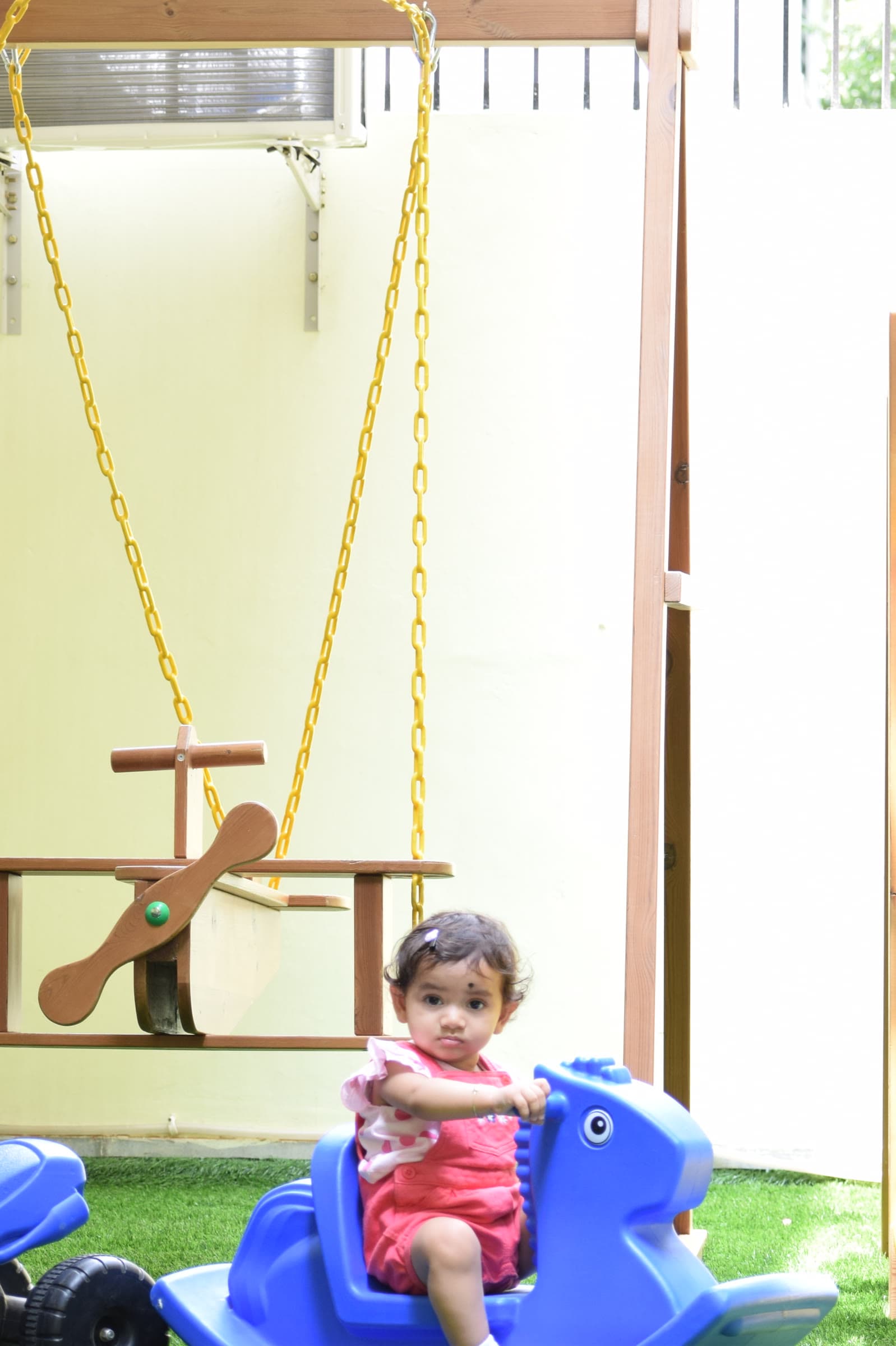 A toddler on a rocking horse on the outdoor terrace, beside a wooden swing and a yellow slide